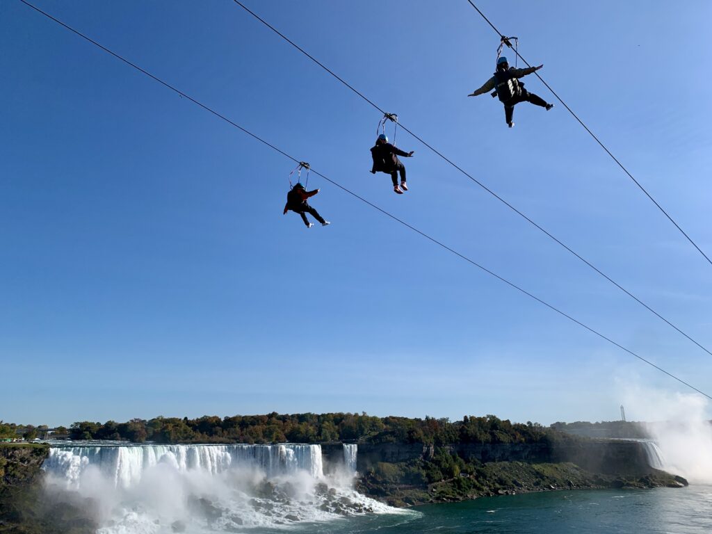 Three people glide on ziplines in clear blue skies in front of the American Falls.