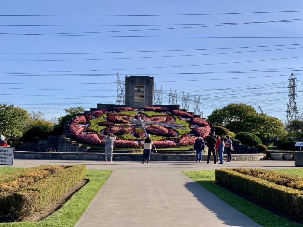 A vibrant floral clock adorned with pink, red, and green flowers, with a group of visitors admiring and taking photos. The clock is framed by neatly trimmed hedges, a blue sky, and utility towers in the distance.