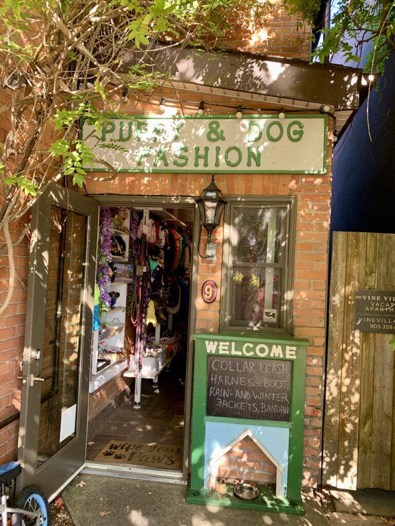 The entrance to the dog shop is in the shade with a small tree in front. It is a small, narrow shop with a green welcome sign out front.