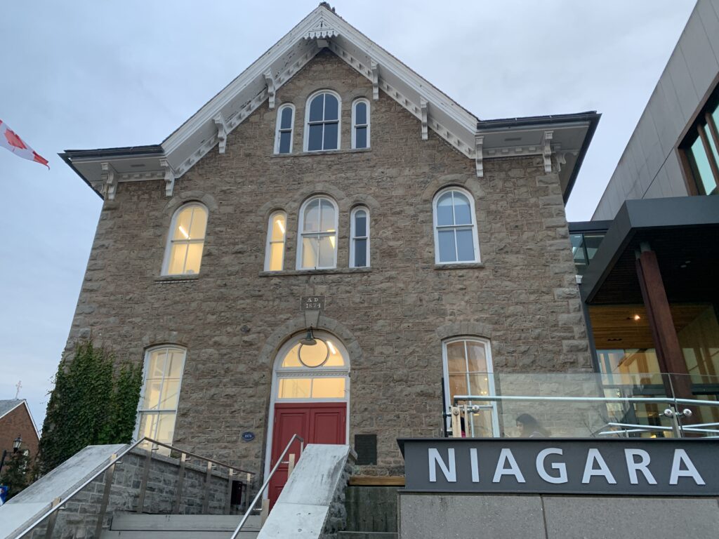 A historic stone building marked "A.D. 1874," with large windows and a bright red door. The modern addition on the right displays the name "Niagara Falls History Museum" on a sleek sign, blending old and new architectural styles.