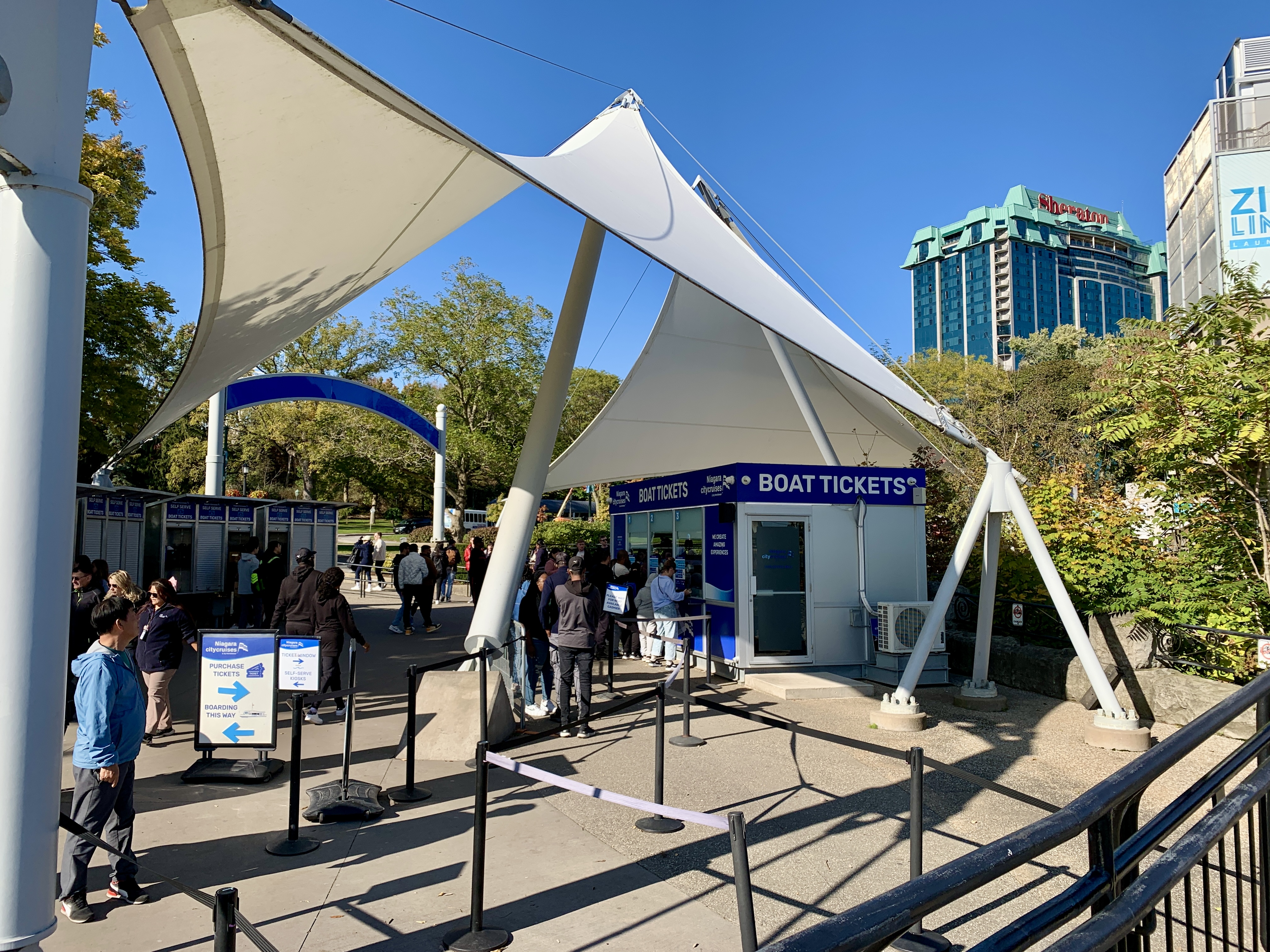 Ticket booth area for Niagara City Cruises, featuring a small white kiosk under large, modern sail-shaped canopy structures. Visitors are seen purchasing tickets and walking around the area, with signs pointing to the boarding area and a backdrop of trees and the Sheraton hotel in the distance under a clear blue sky—a useful thing to look for on a 1 day Niagara Falls itinerary.