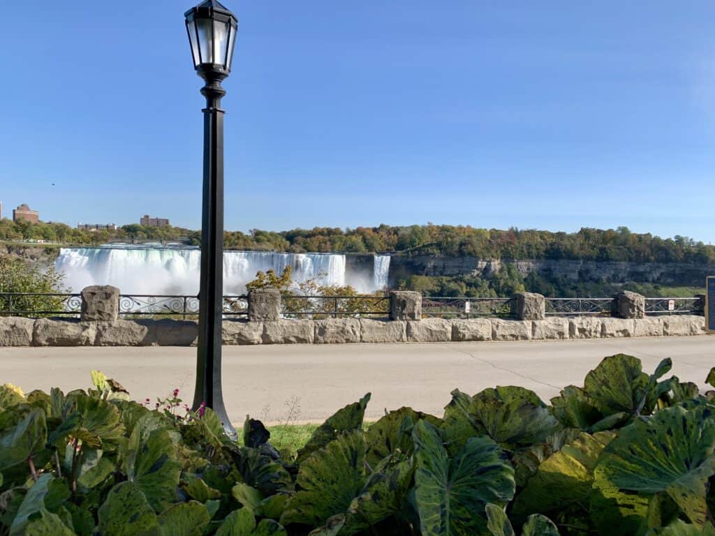 View of the American Falls at Niagara Falls, taken from the Niagara Parkway. A black streetlamp stands in the foreground, with lush green foliage and a stone barrier framing the scene. The waterfall cascades in the distance under a clear blue sky, with autumn-colored trees and buildings visible across the river.