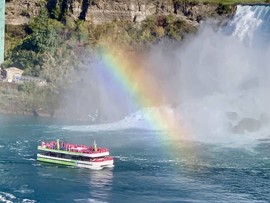 The Niagara City Cruises boat, filled with passengers in red rain ponchos, glides through the blue waters near the base of the American Falls. A vibrant rainbow arches through the mist created by the cascading water, with rocky cliffs and lush greenery in the background—a must-see stop for any 1 day Niagara Falls itinerary.