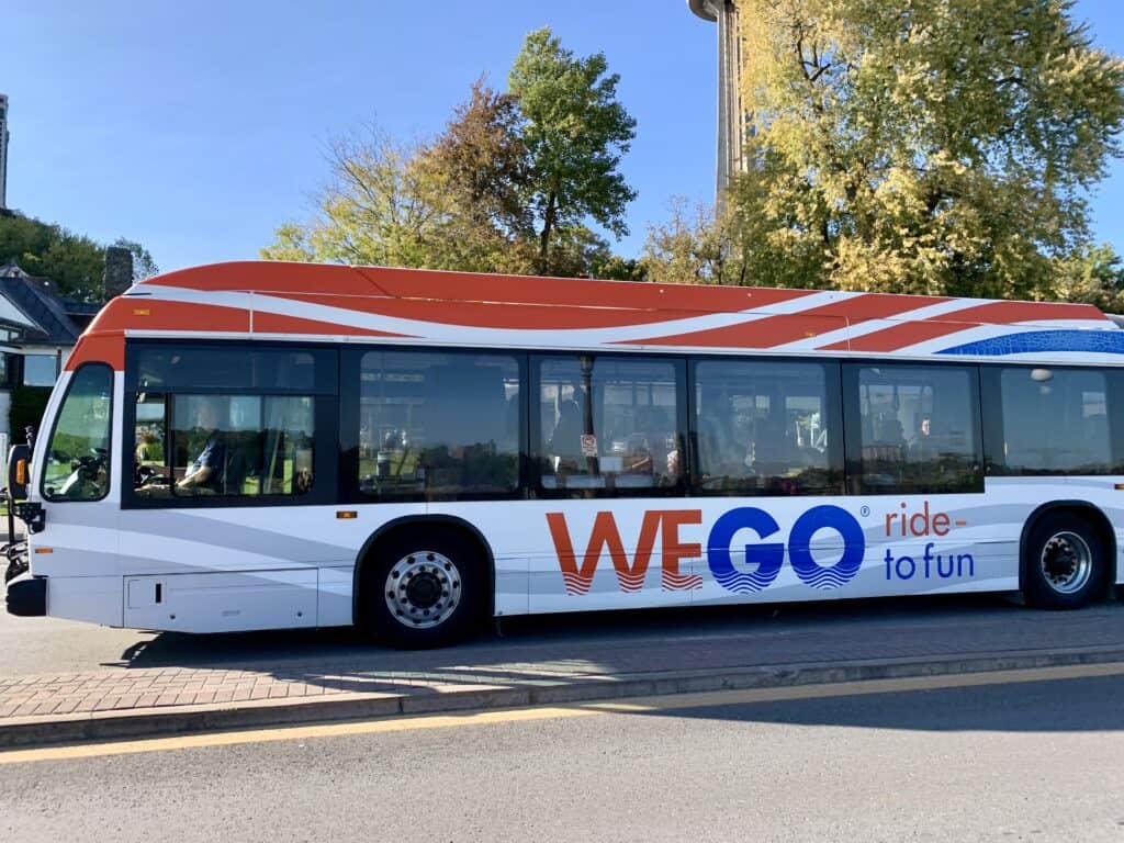 A WEGO bus in Niagara Falls with bold red, white, and blue graphics and the slogan 'ride to fun' displayed on its side. The bus is parked on a sunny day near trees and buildings, with the Skylon Tower visible in the background—a useful thing to look for on a 1 day Niagara Falls itinerary.