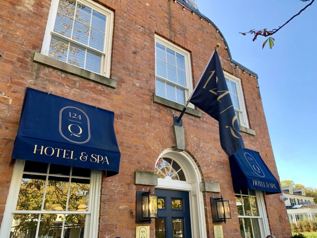 The entrance of 124 Queen Hotel & Spa in Niagara-on-the-Lake, featuring navy-blue awnings and a flag with the hotel's logo. The brick building stands out against a clear sky.