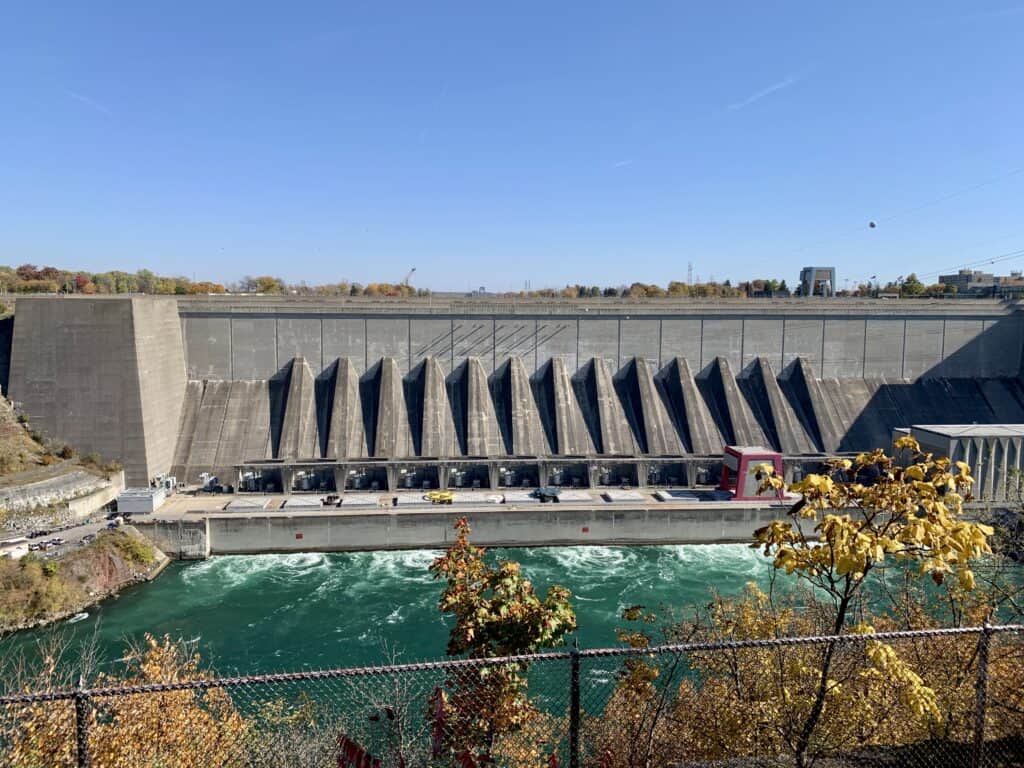 The Robert Moses Niagara Power Plant, a massive concrete hydroelectric facility with distinct triangular structures along its base, sits alongside the rushing turquoise waters of the Niagara River. Autumn trees frame the view, making it an interesting stop on a 1 day Niagara Falls itinerary.