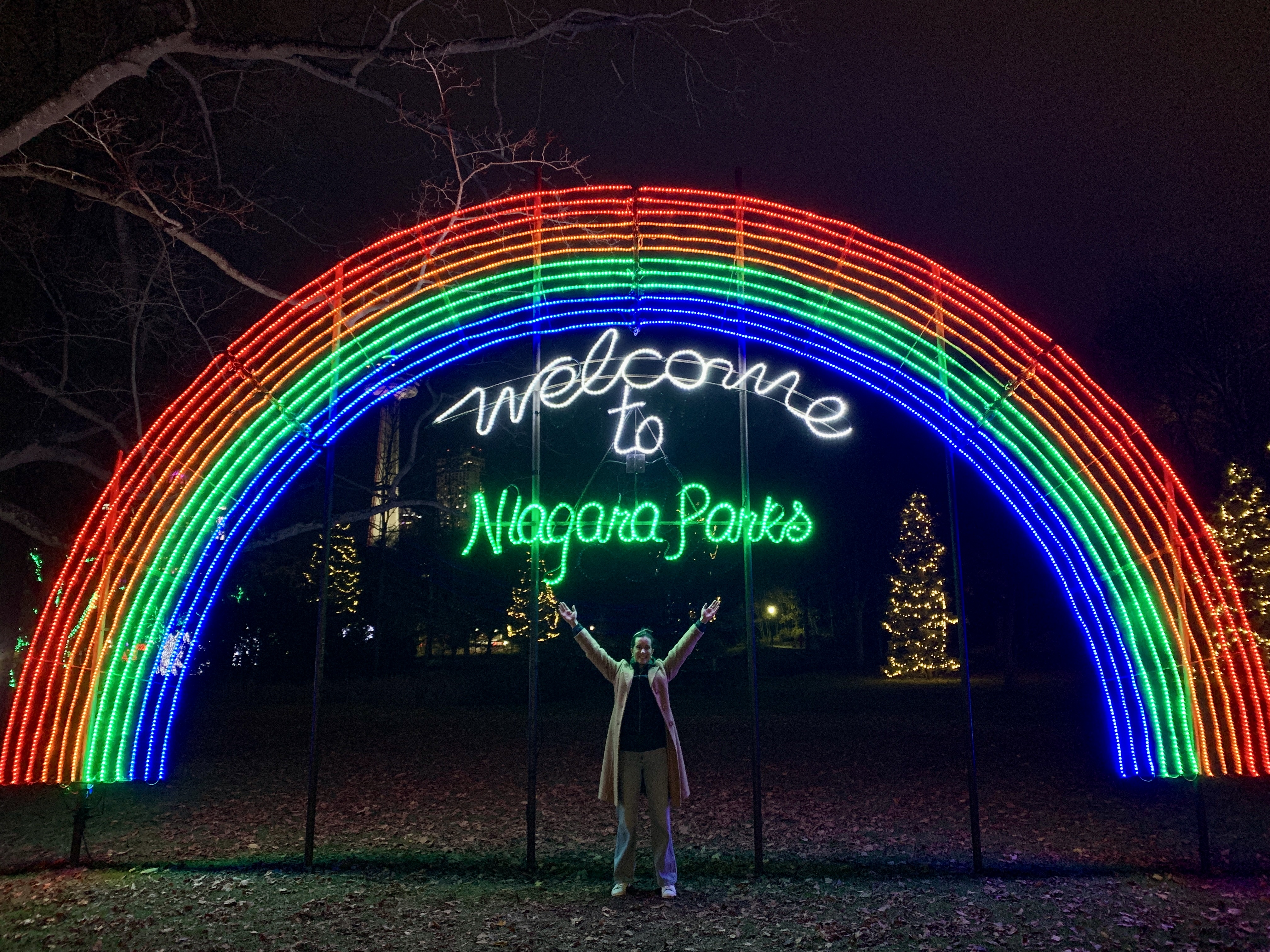 A vibrant rainbow-shaped light display reading "Welcome to Niagara Parks" during the Festival of Lights in Niagara Falls. The author poses joyfully beneath the illuminated arch, surrounded by trees lit with string lights.