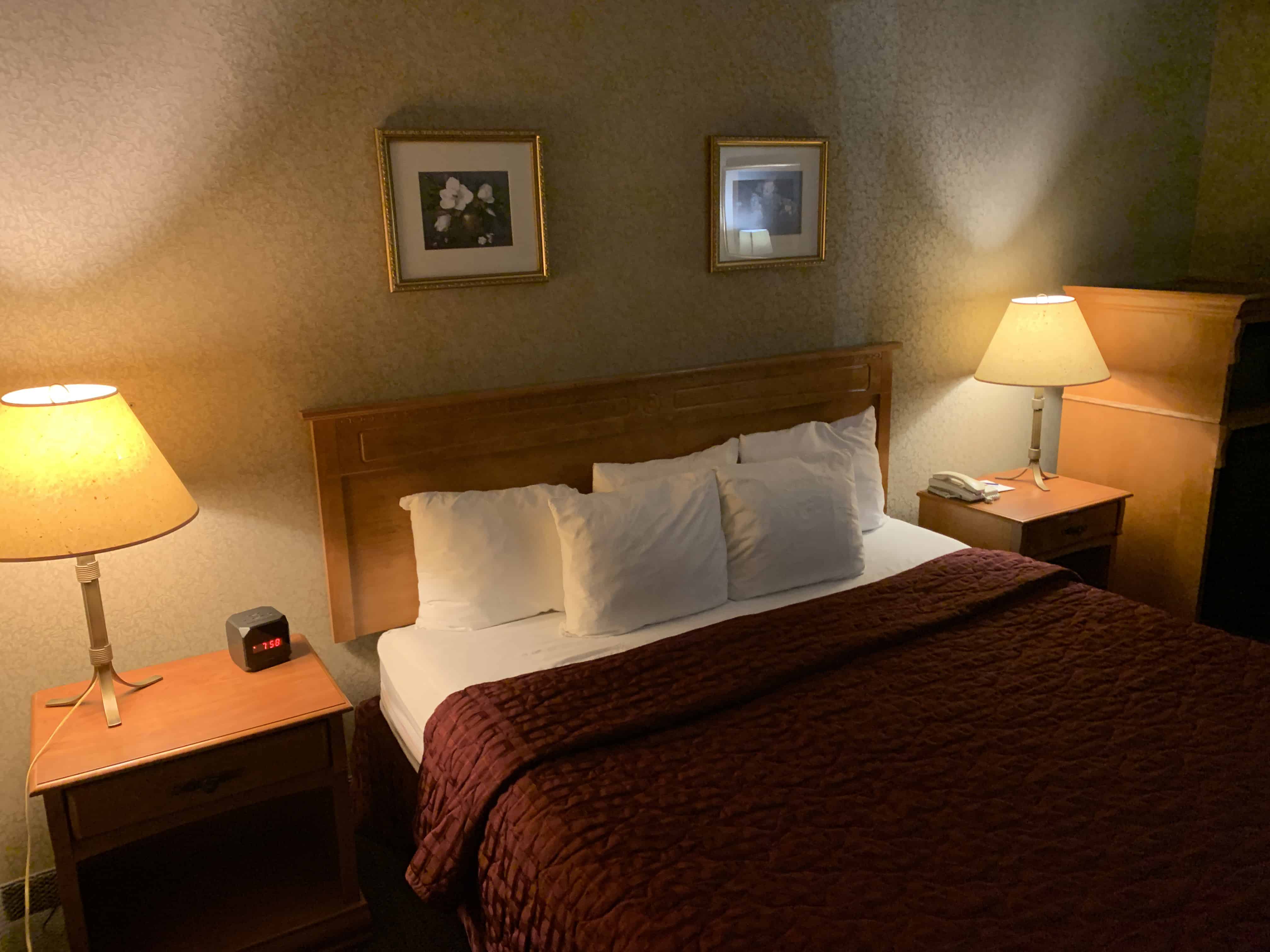 A cozy hotel room at the Comfort Inn featuring a neatly made bed with a burgundy quilt and multiple white pillows. Two wooden nightstands with matching lamps flank the bed, and framed artwork decorates the wallpapered walls—a comfortable accommodation option for a 1 day Niagara Falls itinerary.