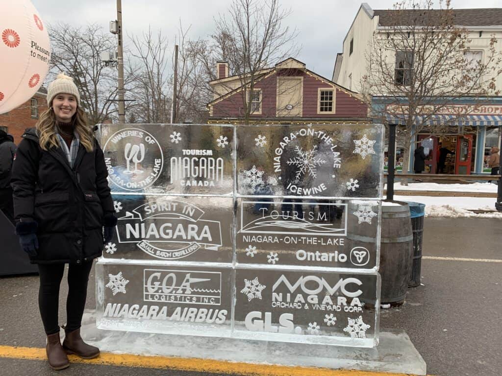 The author stands beside an ice sculpture featuring logos for the Niagara-on-the-Lake Icewine Festival. The sculpture showcases local sponsors and wineries against the backdrop of a lively winter festival scene.