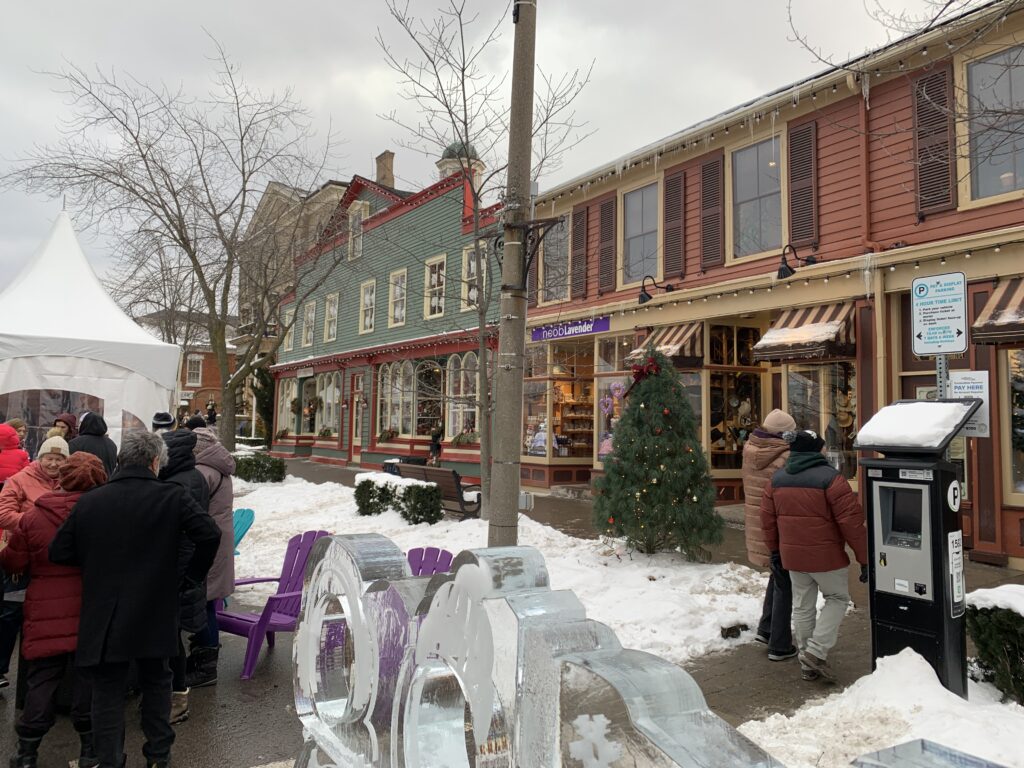 A charming street in downtown Niagara-on-the-Lake during winter, featuring historic storefronts adorned with festive decorations. Visitors in warm coats line up near ice sculptures as part of the Icewine Festival.