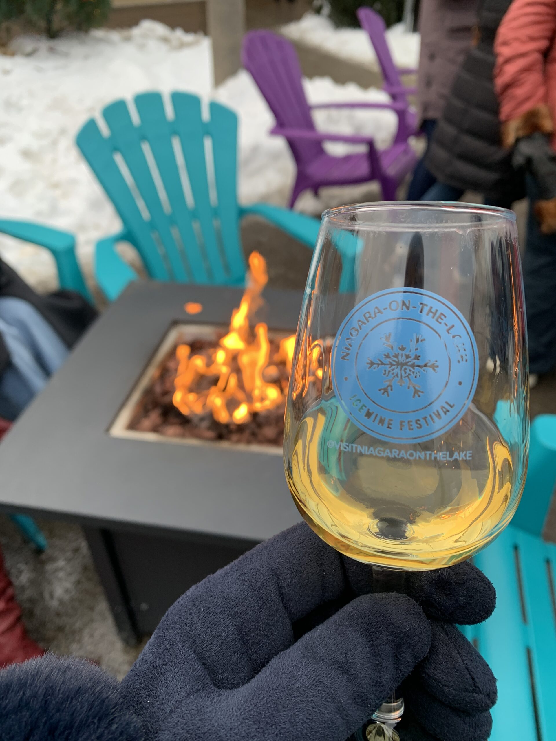 A close-up of a gloved hand holding a glass of golden ice wine at the 2025 Niagara-on-the-Lake Winter Icewine Festival. The wine glass is branded with the festival's logo, and a cozy fire pit with blue Muskoka chairs visible in the background.