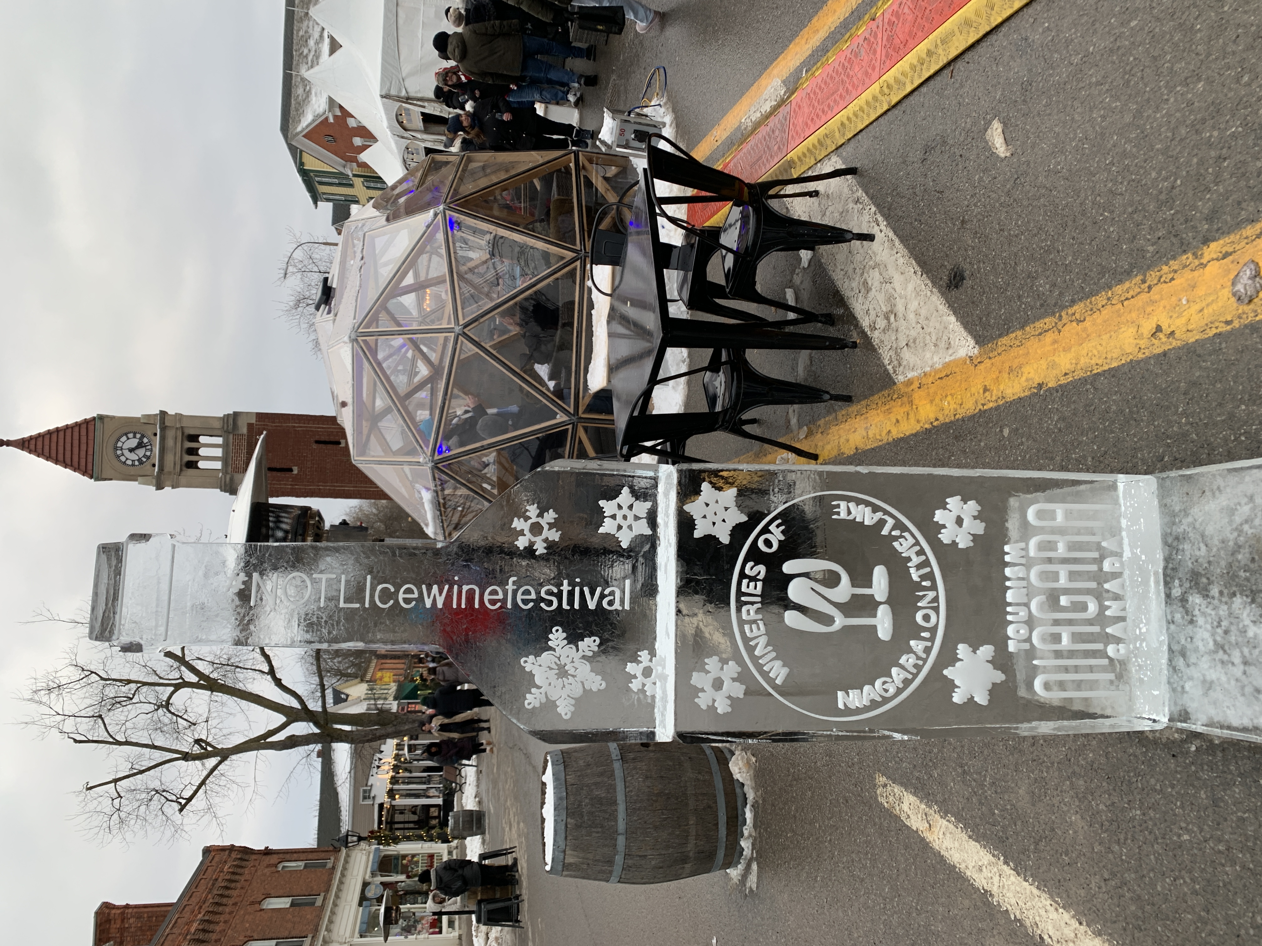 A tall ice sculpture shaped like a wine bottle stands on Queen Street during the 2025 Niagara-on-the-Lake Icewine Festival. It features logos for "Tourism Niagara Canada" and local wineries, with festival-goers and decorative domes in the background.