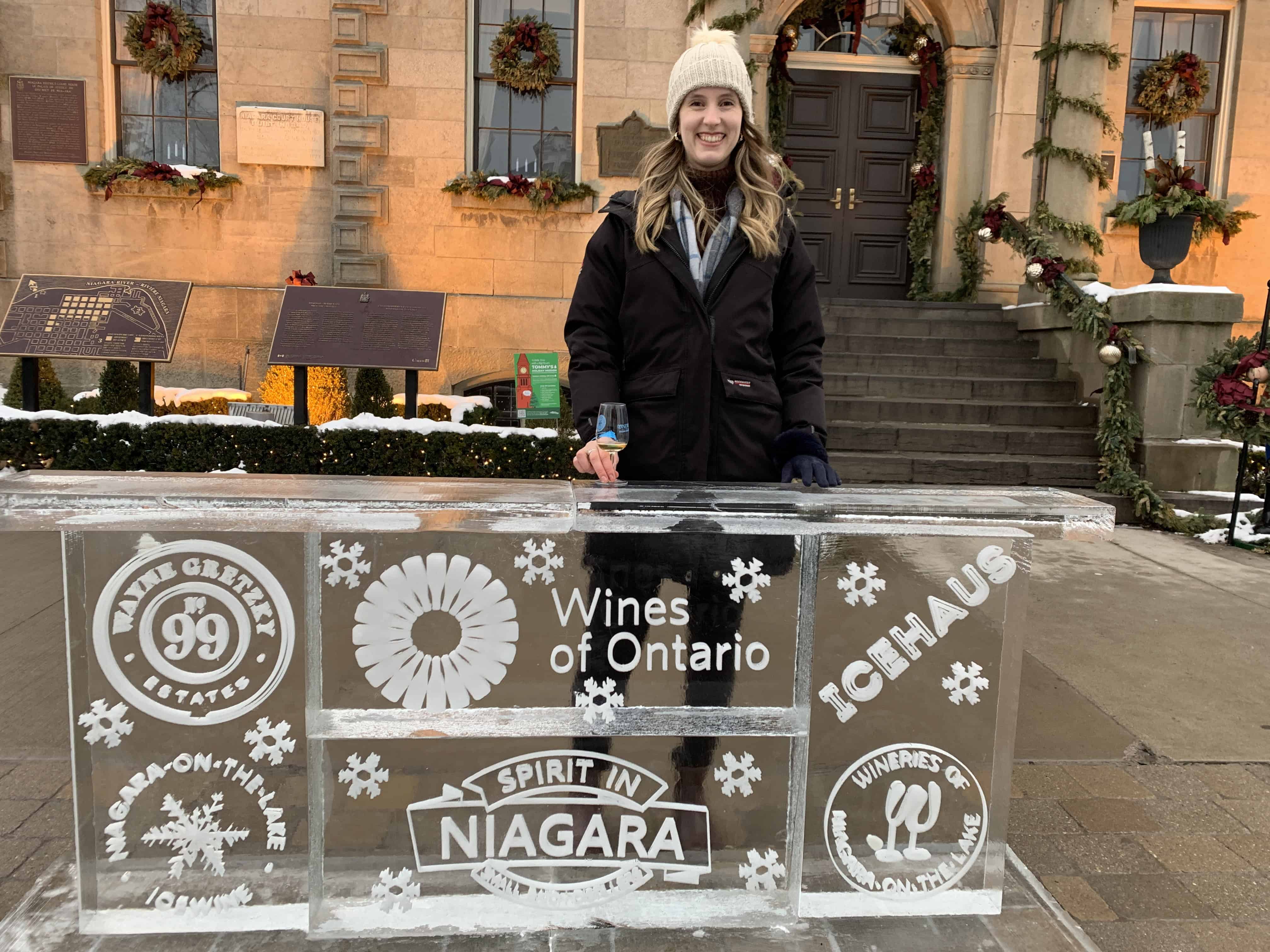 A woman smiles while standing behind an ice sculpture at the 2025 Niagara-on-the-Lake Icewine Festival. The sculpture displays logos for "Wines of Ontario" and other local wineries, with a historic building decorated for winter in the background.