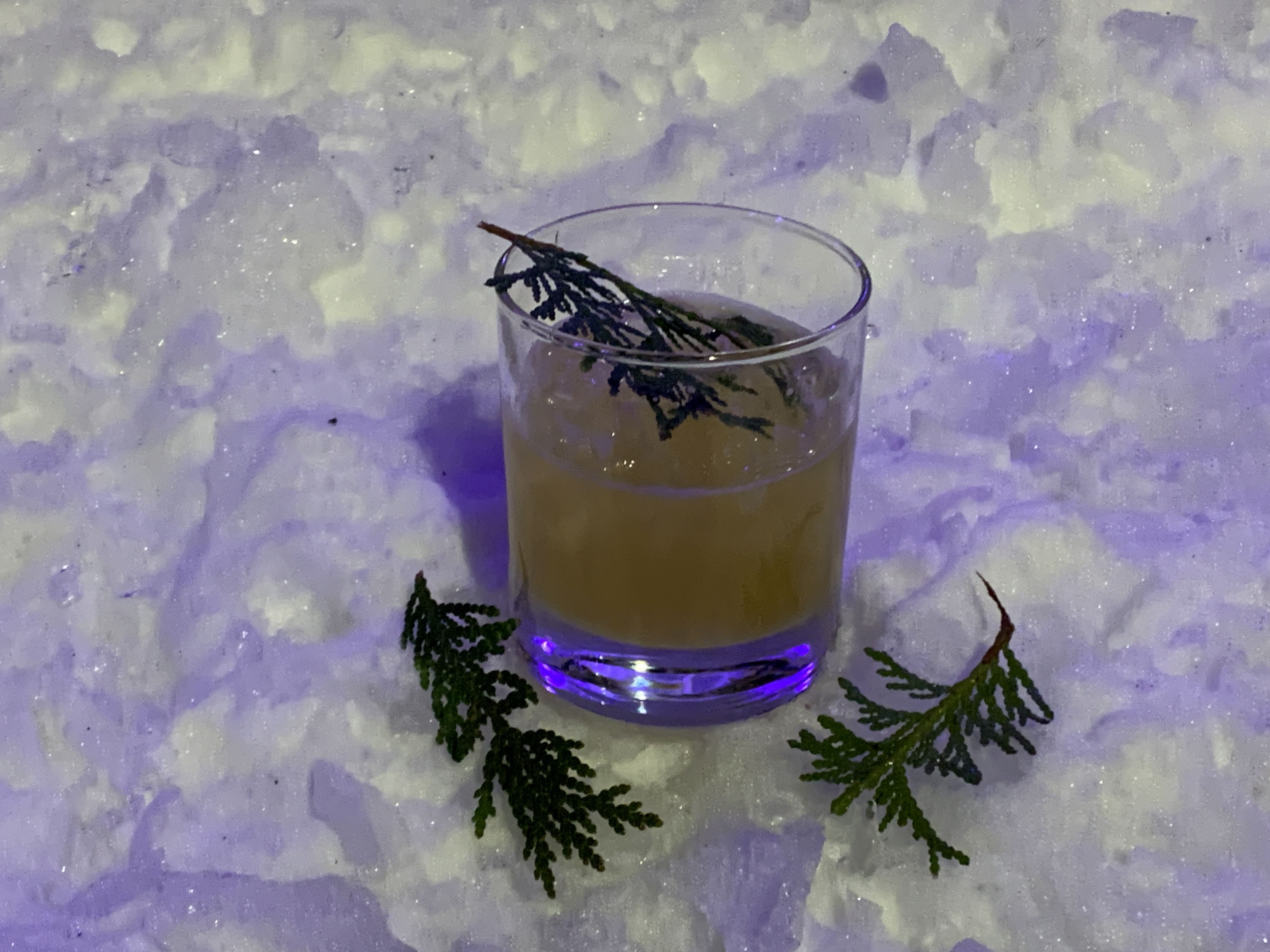 A cedar tea cocktail sits on a snowy surface at the Niagara-on-the-Lake Icewine Festival. The glass is garnished with sprigs of cedar, illuminated by soft purple lighting, capturing a cozy winter aesthetic.