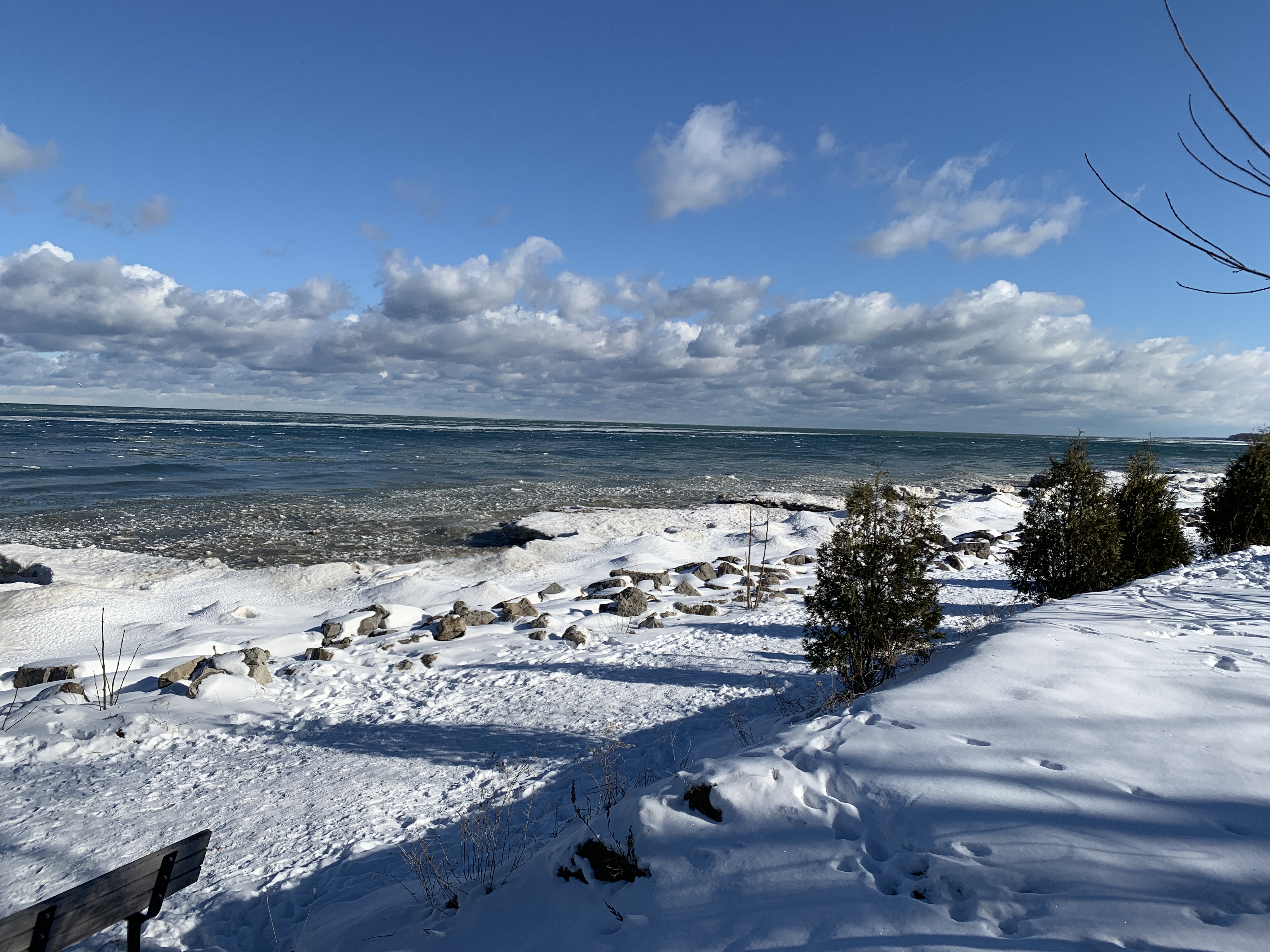 A peaceful walking path along the frozen shore of Lake Ontario in Niagara-on-the-Lake during winter. Snow-covered rocks and small trees line the scenic, icy waterfront.