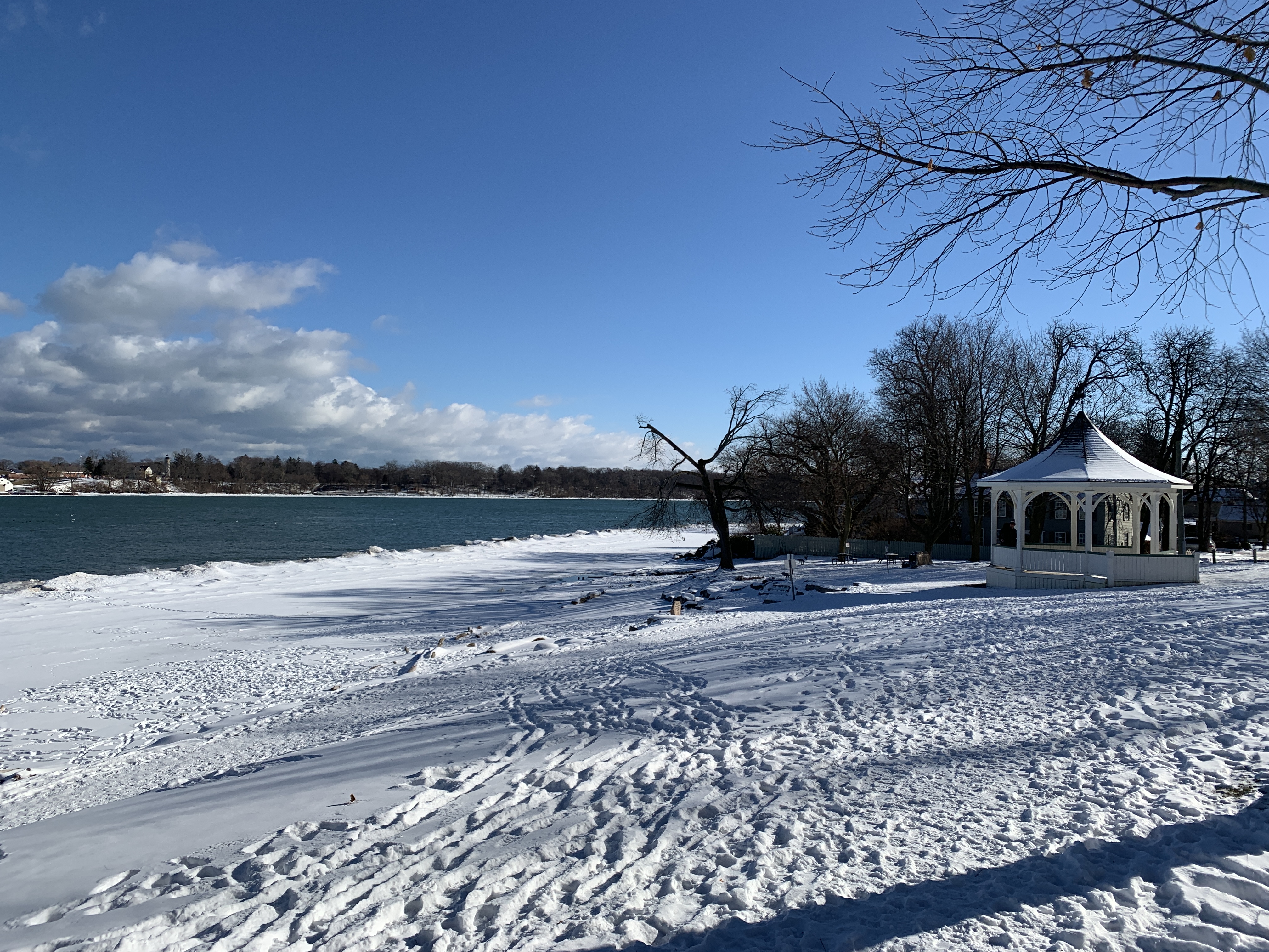 A snowy Queen's Royal Park in Niagara-on-the-Lake during winter, featuring a gazebo near the shoreline of Lake Ontario. Bright sunshine and clear blue skies contrast with the winter landscape.