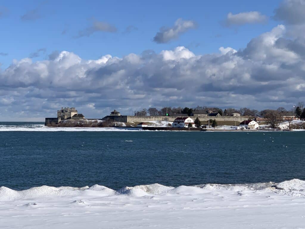 A view across the Niagara River from Niagara-on-the-Lake to Fort Niagara in New York, USA. Snow blankets the ground while the historic fort stands prominently under a partly cloudy sky.