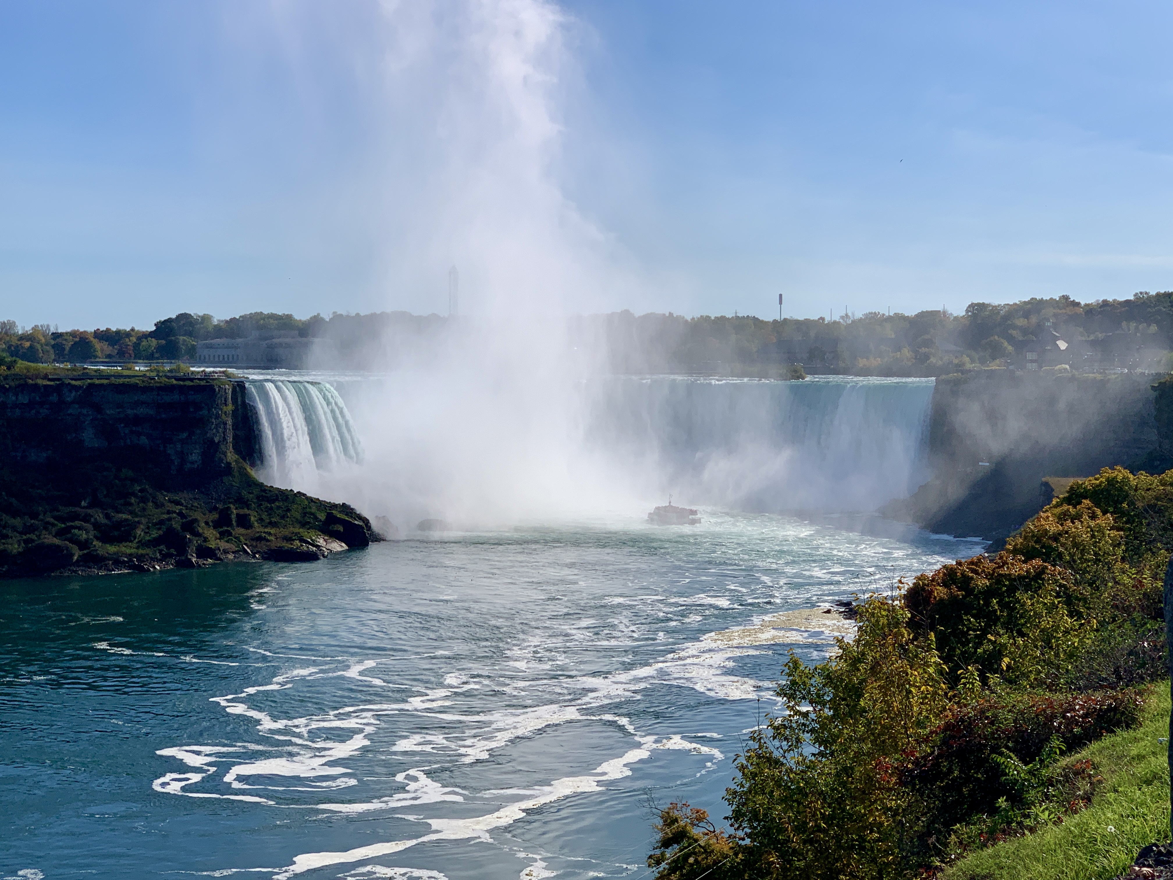 A dramatic shot of Niagara Falls with mist rising high into the blue sky as water thunders over the cliffs. The turquoise river below reflects the sunlight, with a boat barely visible near the base of the falls and lush greenery in the foreground. Niagara Falls is a great idea for day trips from Toronto.