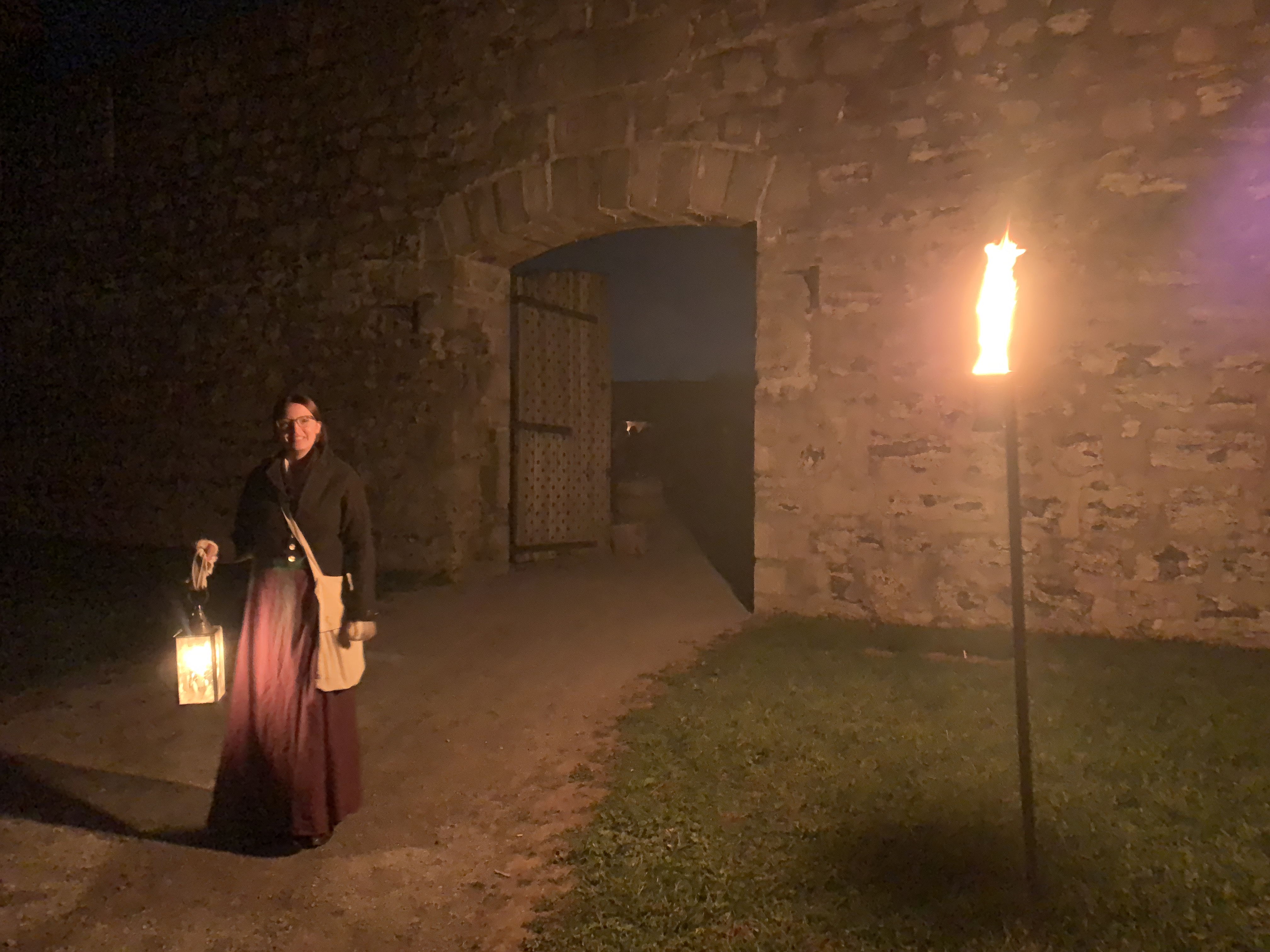 A nighttime scene at Old Fort Erie, with a guide dressed in period attire holding a glowing lantern. The dimly lit fort gate and a torch in the foreground create a haunting atmosphere. Going on the ghost tour is a great idea for day trips from Toronto.