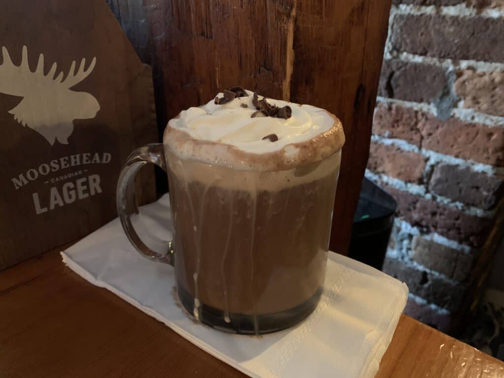 A rich hot chocolate topped with whipped cream and chocolate shavings, served in a clear glass mug at the Olde Angel Inn in Niagara-on-the-Lake. The rustic wooden and brick backdrop adds to the cozy, historic ambiance.