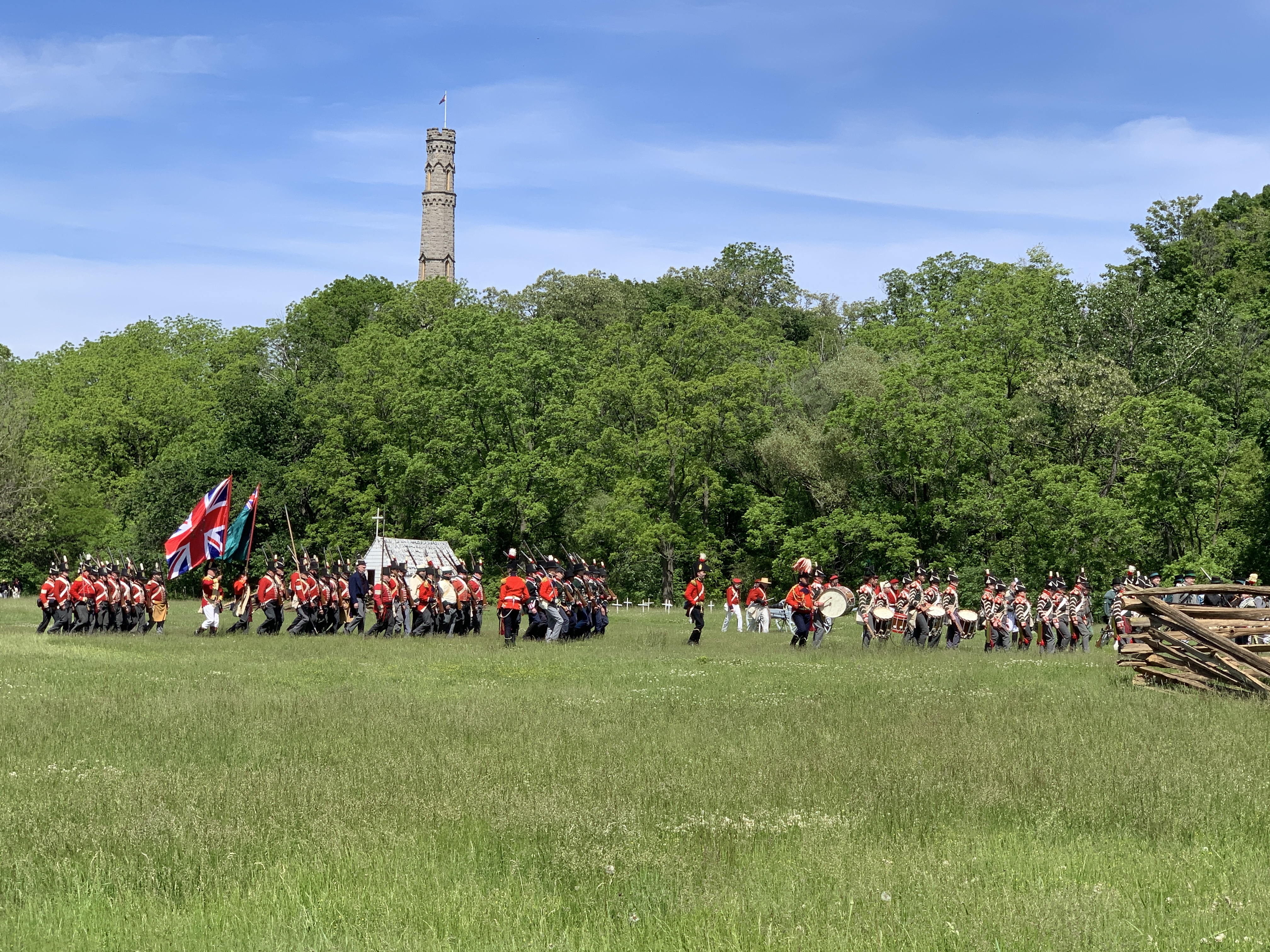 A super fun day trip from Toronto is the reenactment of the Battle of Stoney Creek, featuring soldiers in red and white uniforms marching in formation with flags waving. A historic stone monument rises in the background amid lush greenery under a clear blue sky.