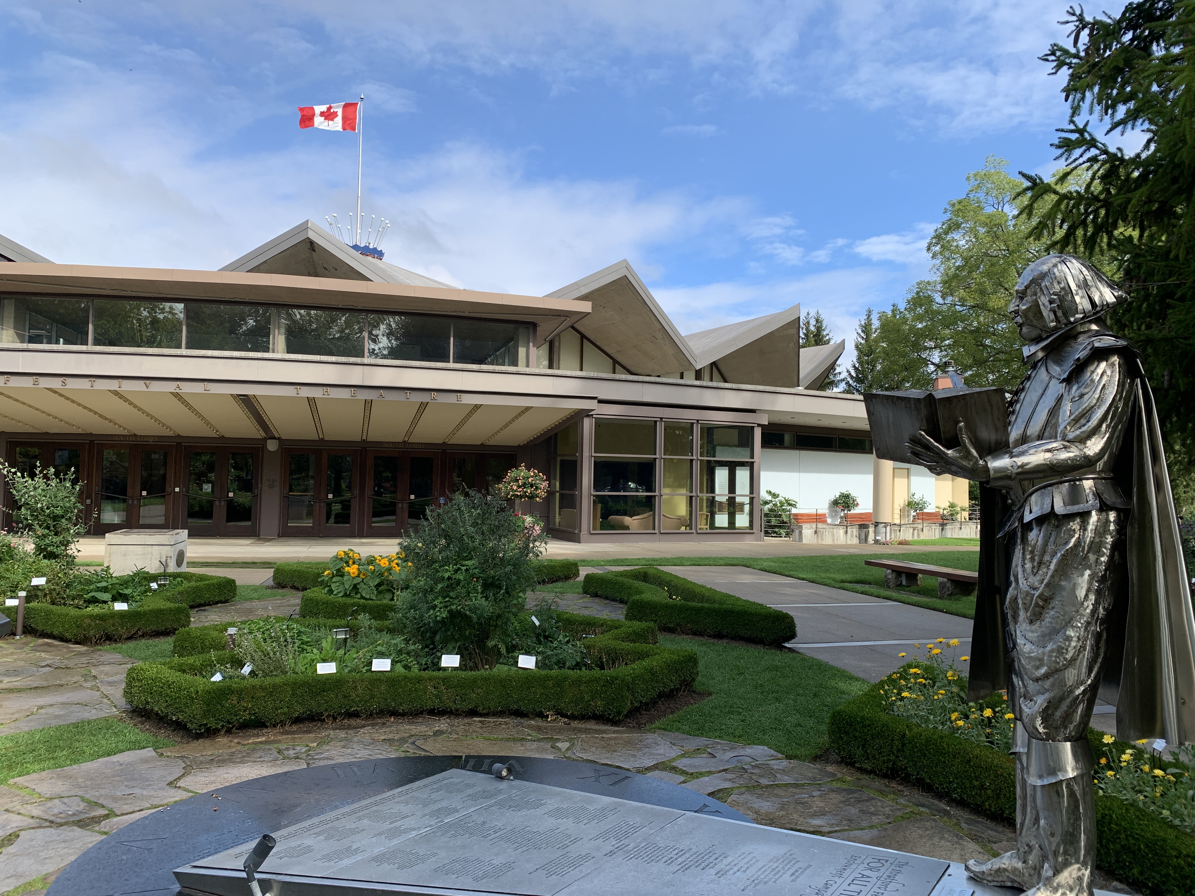 The Festival Theatre in Stratford, with its distinctive angular rooflines, surrounded by manicured gardens and a statue of Shakespeare holding an open book. A Canadian flag flies prominently against a blue sky.