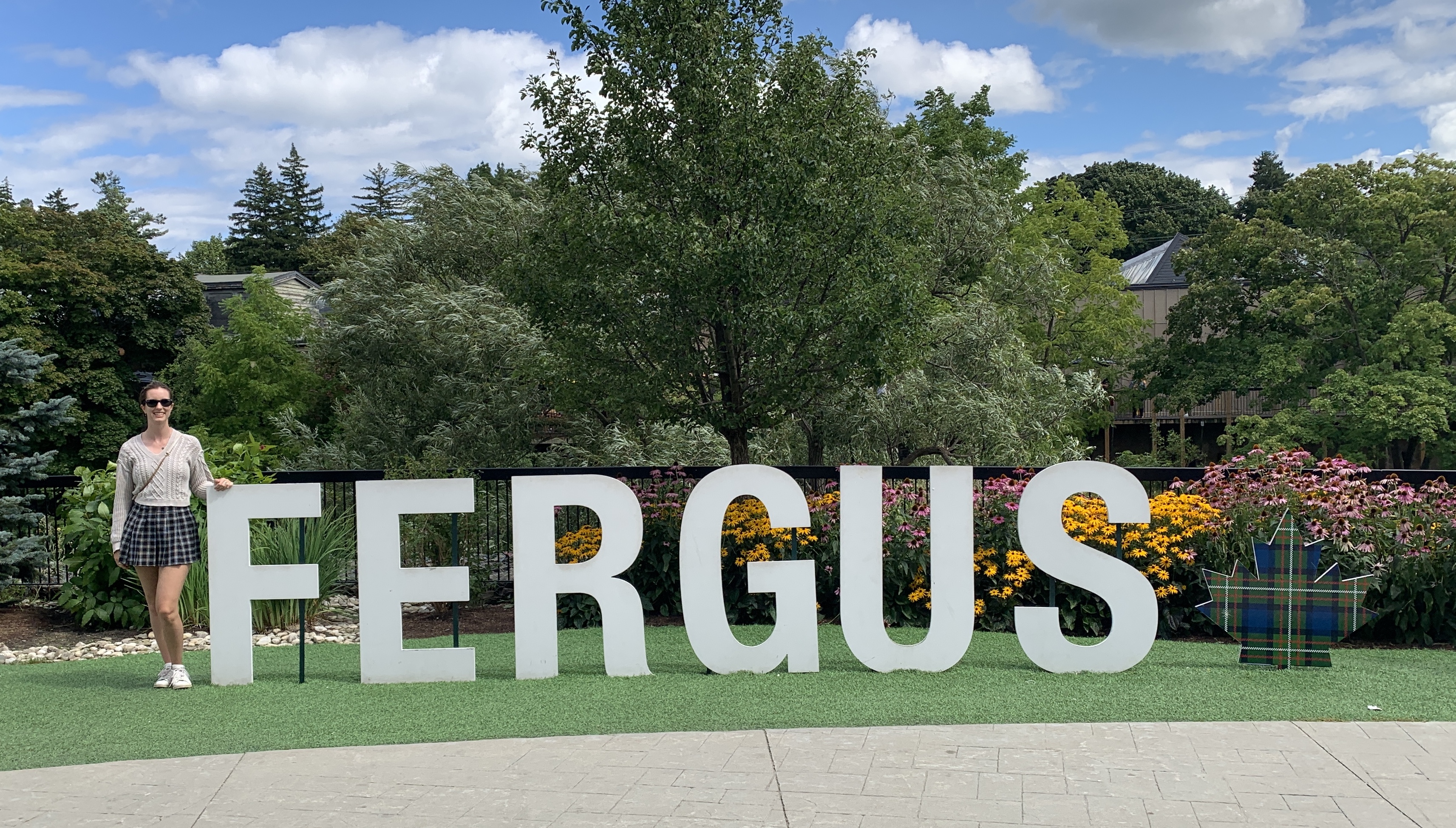 The author standing next to a large "FERGUS" sign on a grassy area, surrounded by colourful flowers and trees under a bright blue sky. The sign includes a plaid maple leaf symbol, adding a touch of Canadian-Scottish flair. Attending the Highland games is a great idea for day trips from Toronto.