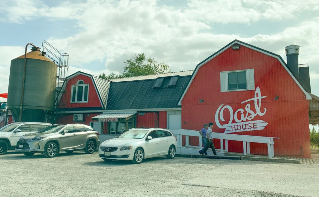 Oast Brewery in Niagara-on-the-Lake, a vibrant red barn-style building with white lettering and silo. The sunny day and parked cars add a welcoming rural charm.